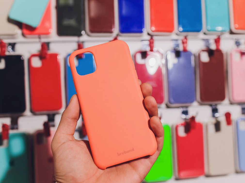 Apple Handyhülle aus Silikon von Breband in Orange, gehalten vor einer Wand mit verschiedenen bunten Handyhüllen.