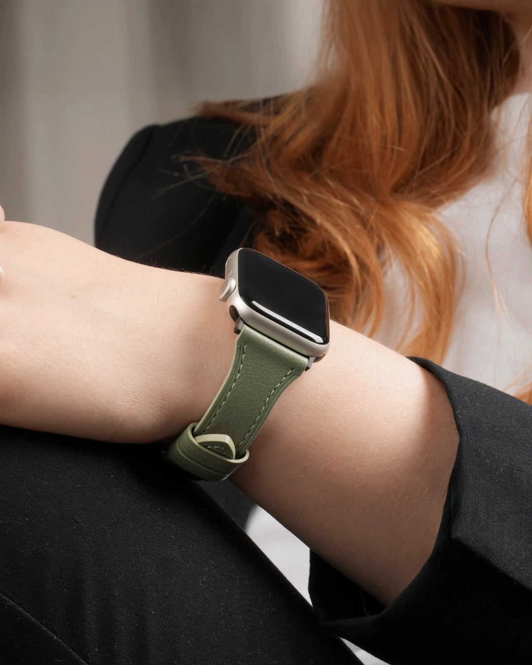 Apple Watch mit grünem Lederarmband am Handgelenk einer Person mit rotem Haar und schwarzem Blazer